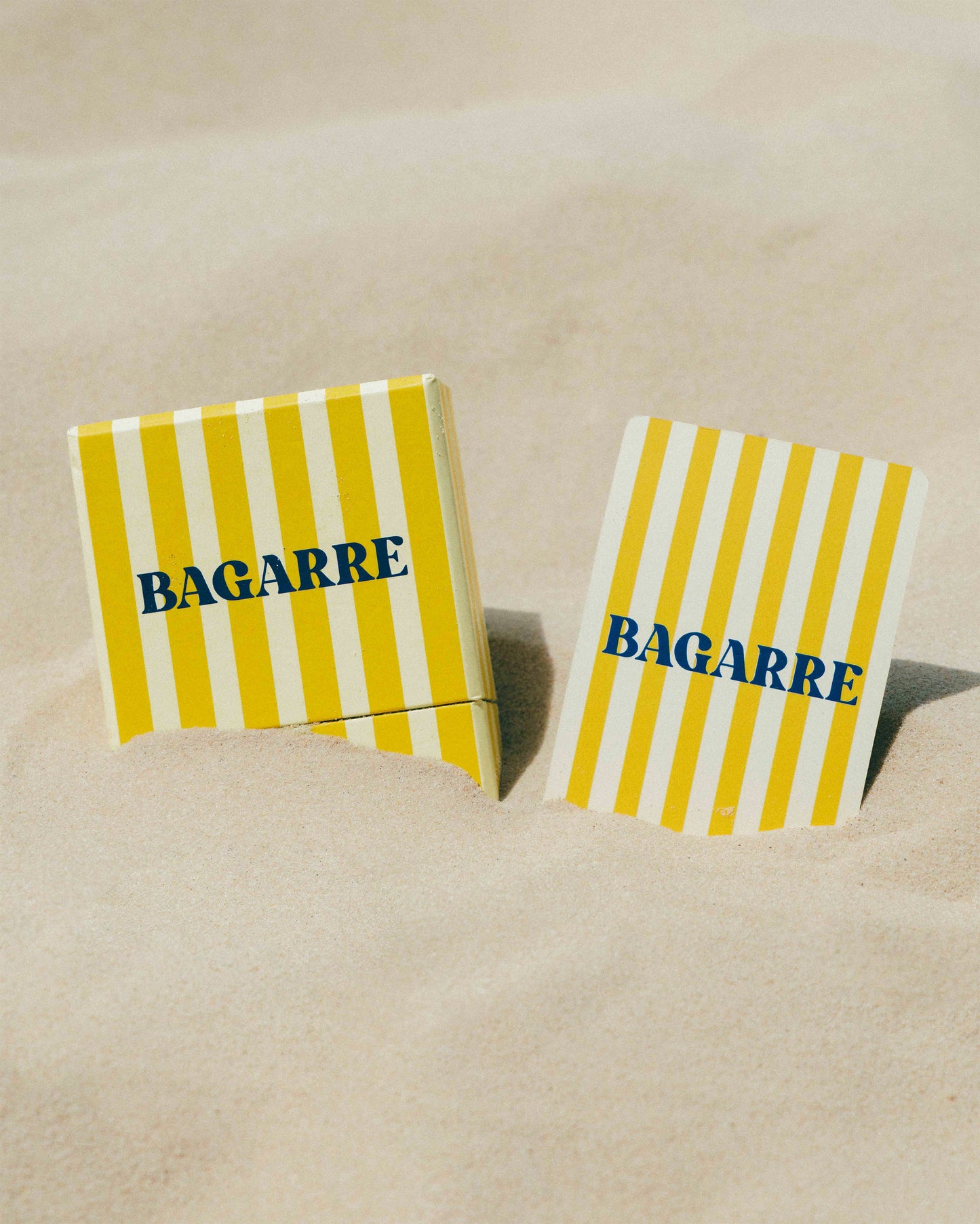 Two yellow and white striped coasters with 'BAGARRE' text on a sandy background
